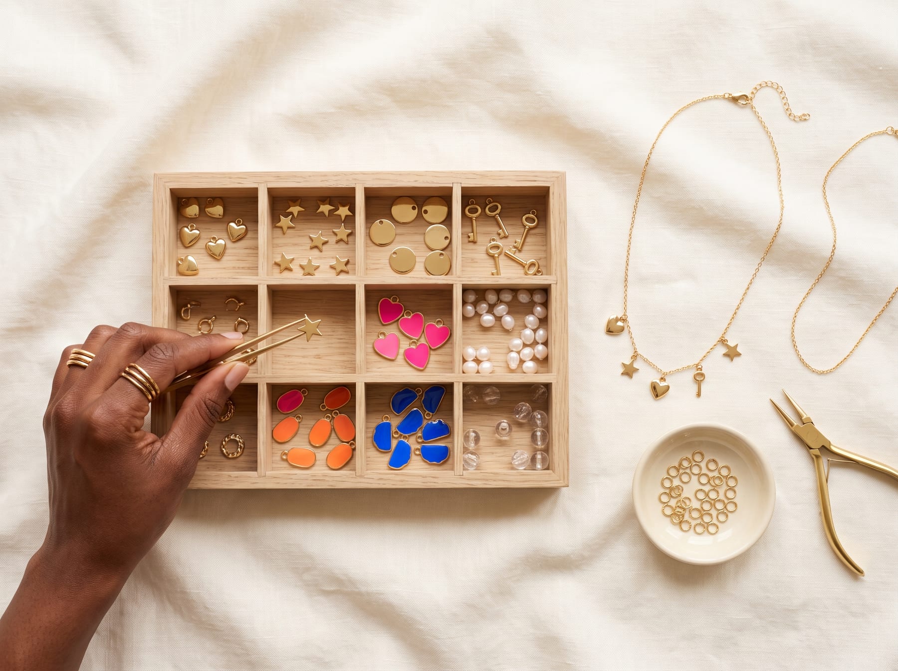 A hand lifting a small gold star charm with tweezers from a compartment tray of gold, pearl and enamel charms, beside a finished gold charm necklace