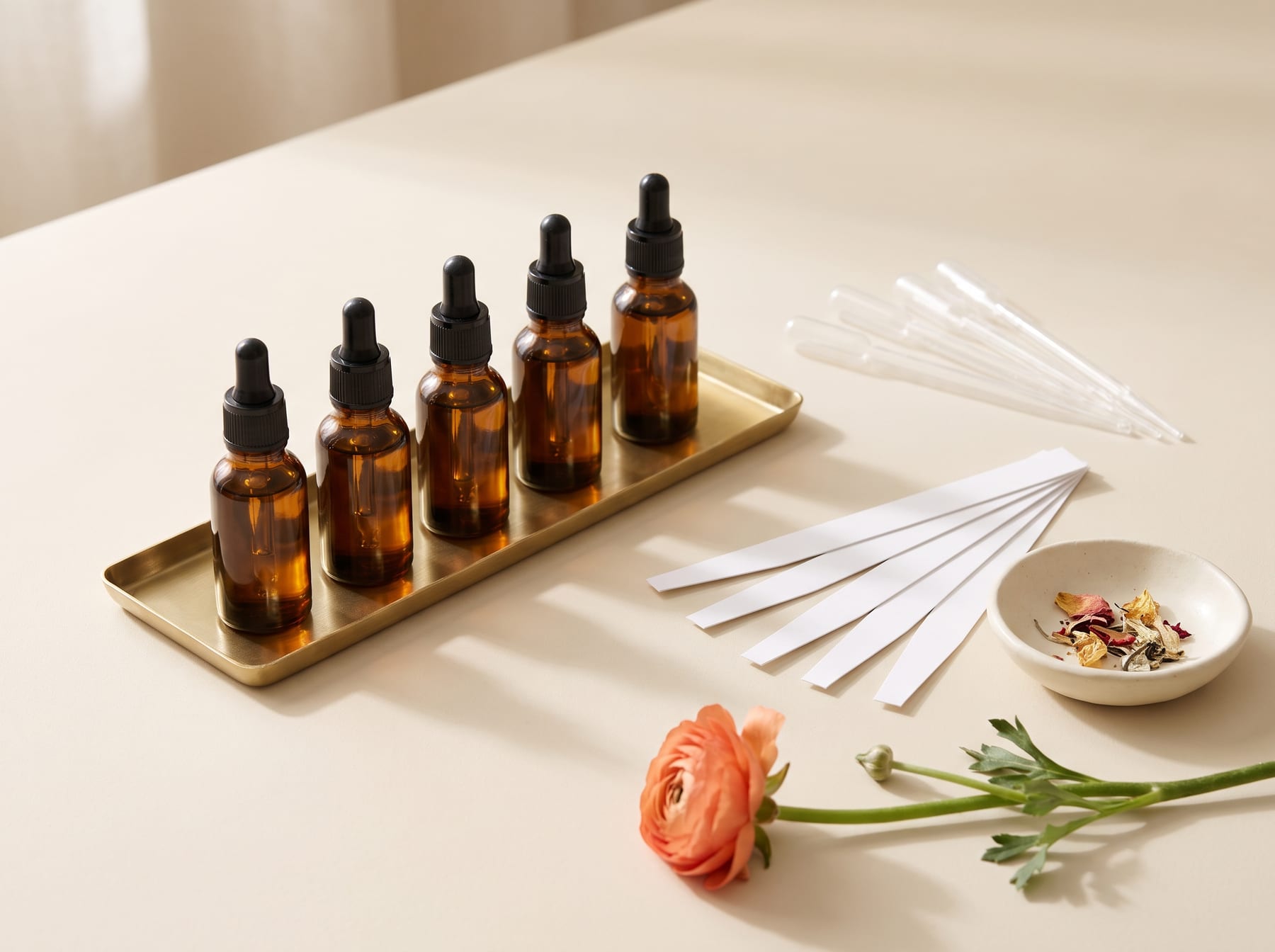 Amber glass dropper bottles lined up on a brass tray beside glass pipettes, paper scent blotters, a dish of dried florals and a single coral ranunculus