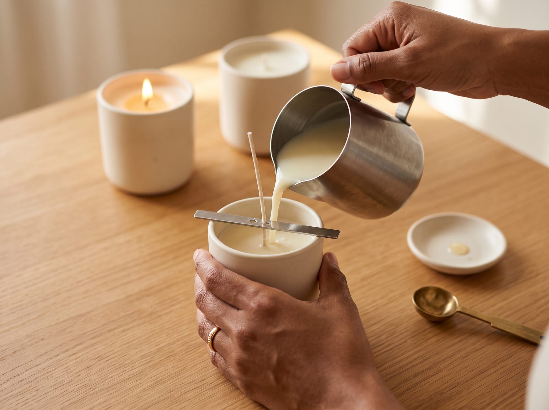 Hands pouring warm cream-colored wax into a speckled ceramic candle vessel, with a lit finished candle alongside